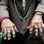 Close-up of a person's hands wearing multiple rings, a brown blazer, and a necklace.